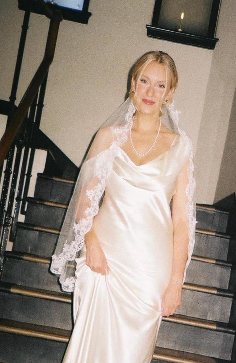 K&ouml;nnte beinhalten: Eine Braut in einem wei&szlig;en Satin-Brautkleid mit Wasserfallausschnitt und einem zarten, mit Spitze besetzten Schleier. Sie tr&auml;gt eine Perlenkette und steht auf einer Treppe mit dunklen Holzgel&auml;ndern.