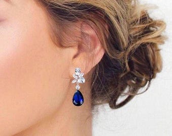 May include: A silver-toned drop earring featuring a cluster of clear, marquise-cut and round-cut stones at the top, leading to a single, large, deep blue, pear-shaped stone at the bottom. The earring is secured with a post and back mechanism.