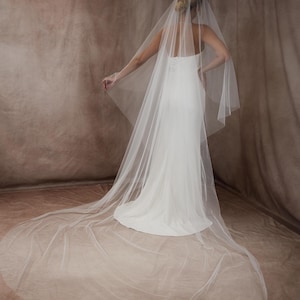 Op de afbeelding: Een witte trouwjurk met een lange sleep en een doorschijnende sluier. De sluier valt over de rug en creëert een vloeiend effect. De jurk heeft een eenvoudig, elegant ontwerp, perfect voor een bruiloft.