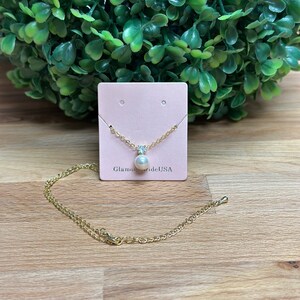 May include: A gold chain necklace with a single pearl pendant and a small clear crystal accent. The necklace is displayed on a pink card with the text "GlamourBrideUSA".