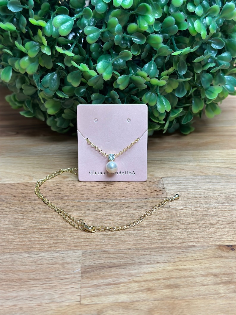 May include: A gold chain necklace with a single pearl pendant and a small clear crystal accent. The necklace is displayed on a pink card with the text "GlamourBrideUSA".