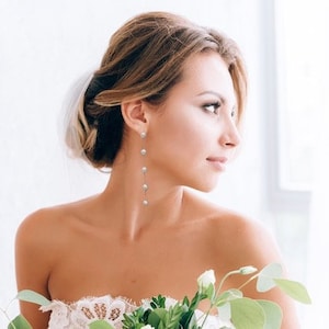 May include: A woman with her hair styled up in a bun wears long pearl drop earrings. The earrings are silver and have five pearls each.