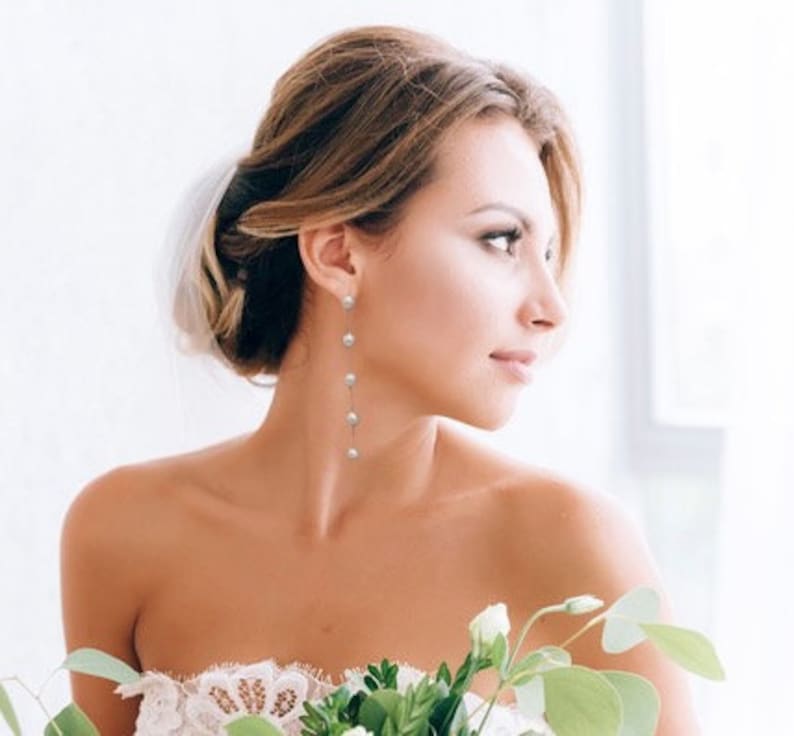 May include: A woman with her hair styled up in a bun wears long pearl drop earrings. The earrings are silver and have five pearls each.