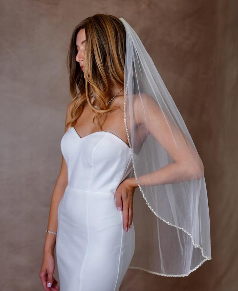 May include: A sheer white bridal veil featuring a delicate beaded edge trim. The veil is made of fine tulle fabric and is draped over the head, extending down the back. It is paired with a simple, strapless white gown with a sweetheart neckline.