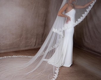 May include: A white wedding veil with a delicate lace trim. The veil is long, flowing, and extends to the floor. The bride is wearing a white strapless gown. The background is a neutral brown.