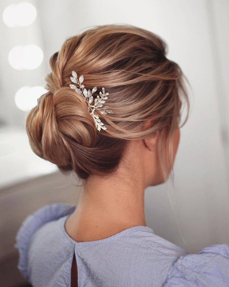 May include: A woman with light brown hair styled in a low bun with a silver and white hair comb. The comb has a leaf design with small white beads.