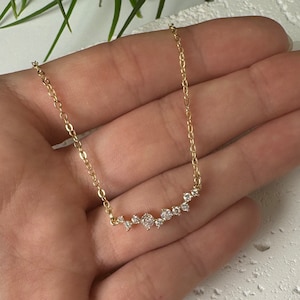 May include: A gold-toned chain necklace featuring a horizontal bar pendant composed of a cluster of small, clear, round-cut stones arranged in a graduated, asymmetrical pattern. The delicate metal chain connects to each end of the stone-set bar.