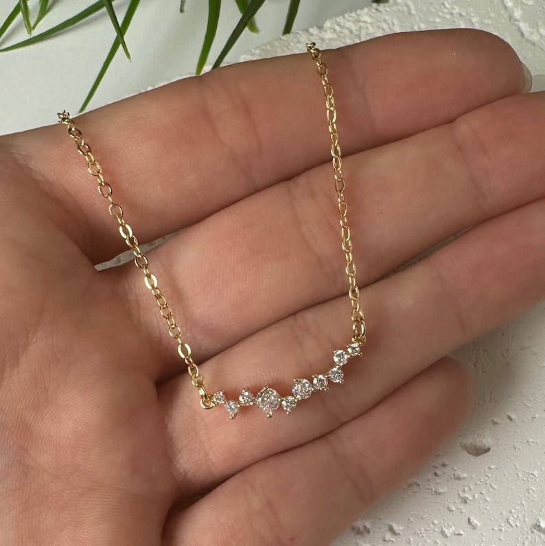 May include: A gold-toned chain necklace featuring a horizontal bar pendant composed of a cluster of small, clear, round-cut stones arranged in a graduated, asymmetrical pattern. The delicate metal chain connects to each end of the stone-set bar.