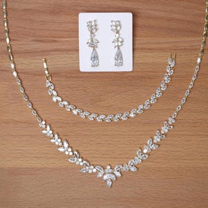 May include: A gold-toned jewelry set featuring a necklace, bracelet, and earrings. The necklace and bracelet have a floral design with clear, sparkling stones. The earrings are teardrop-shaped with matching stones. The set is displayed on a wooden surface.