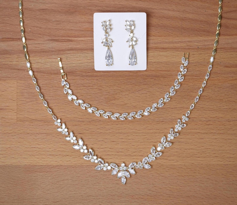 May include: A gold-toned jewelry set featuring a necklace, bracelet, and earrings. The necklace and bracelet have a floral design with clear, sparkling stones. The earrings are teardrop-shaped with matching stones. The set is displayed on a wooden surface.