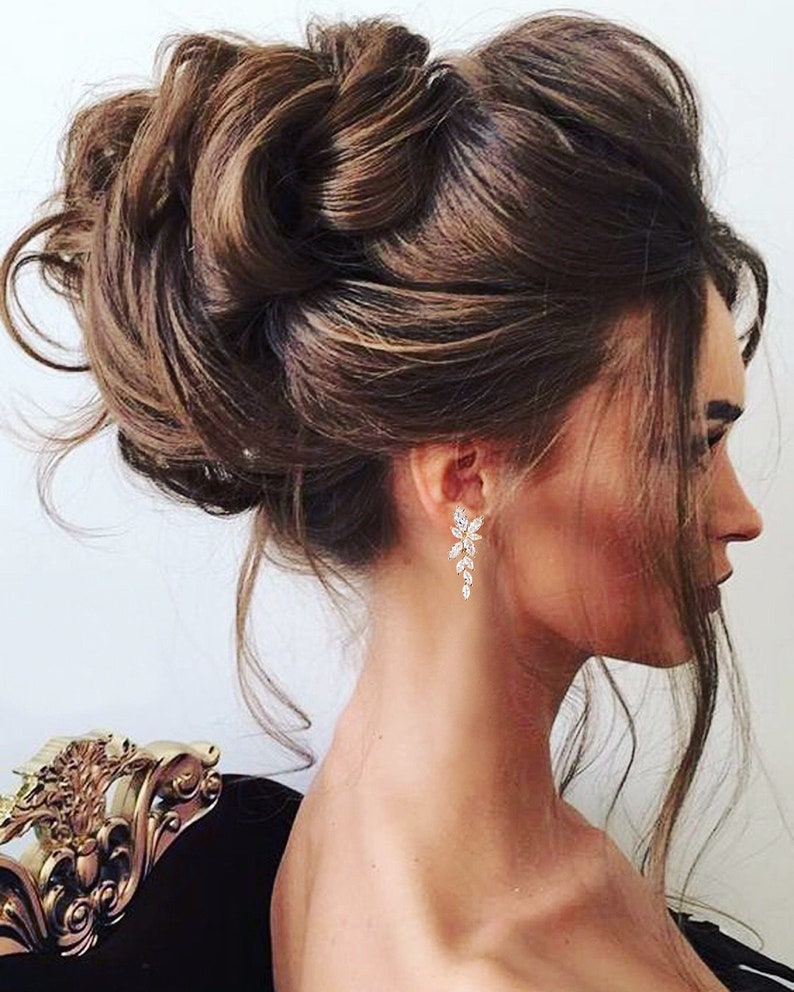 May include: A woman with long brown hair styled in a high bun, wearing long, dangling earrings with clear crystal stones.