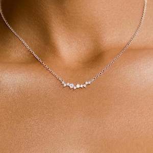 May include: A delicate silver-toned chain necklace featuring a central cluster of seven small, round, clear stones arranged in a horizontal, asymmetrical arc. The stones vary slightly in size, with the largest stone positioned near the center.