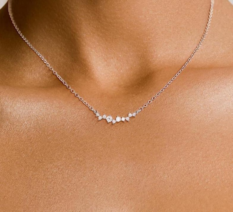 May include: A delicate silver-toned chain necklace featuring a central cluster of seven small, round, clear stones arranged in a horizontal, asymmetrical arc. The stones vary slightly in size, with the largest stone positioned near the center.