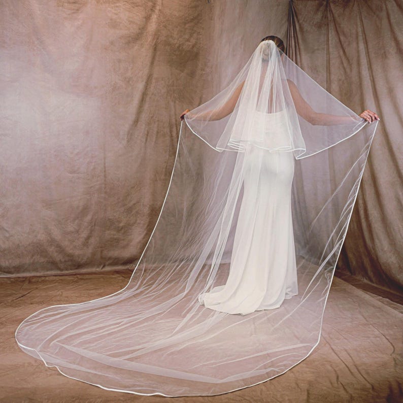 May include: A bride in a white wedding dress stands with arms outstretched, showcasing a long, sheer, white veil with a delicate white trim. The veil cascades down, creating a flowing effect. The background is a neutral brown.
