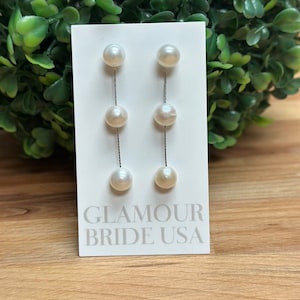 May include: A pair of pearl drop earrings with a silver chain. The earrings are on a white card with the text "Glamour Bride USA".