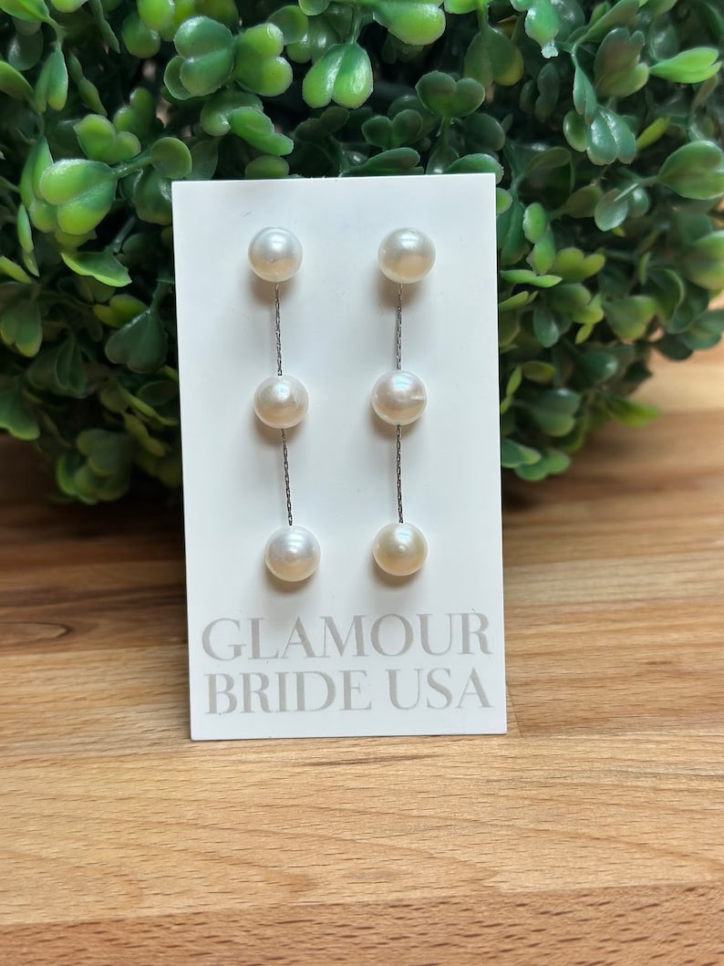 May include: A pair of pearl drop earrings with a silver chain. The earrings are on a white card with the text "Glamour Bride USA".