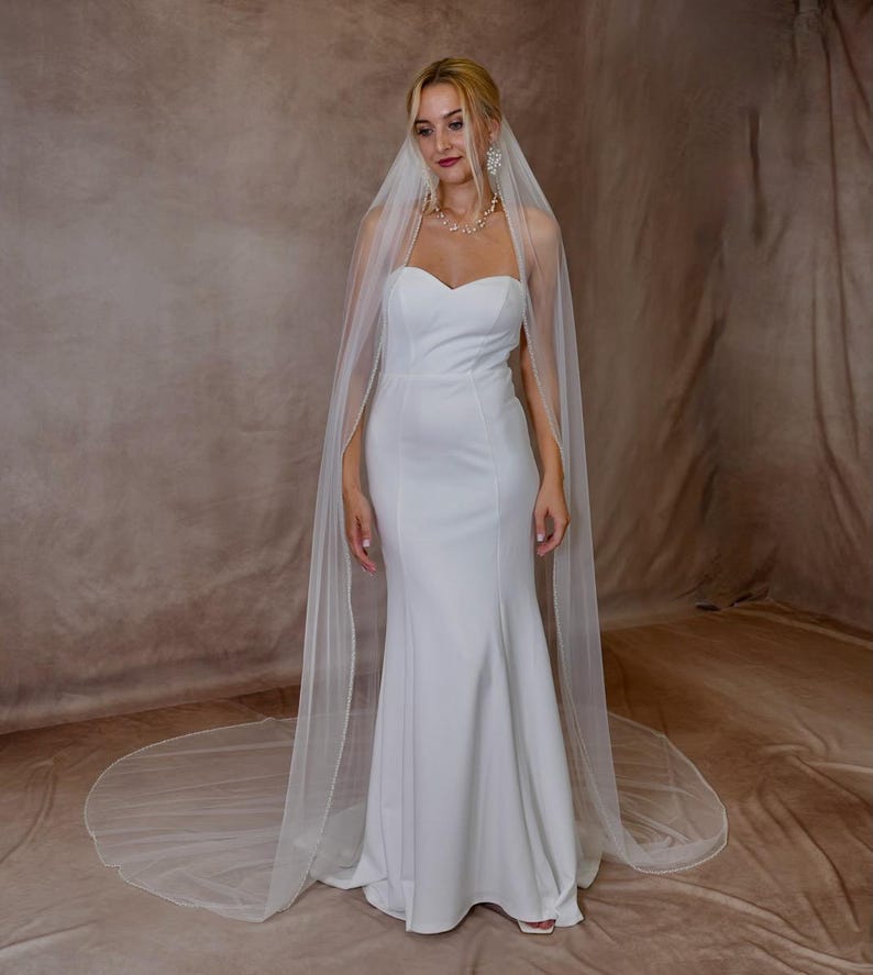 May include: A floor-length white strapless gown with a sweetheart neckline and a fitted bodice that flares at the skirt. A long, sheer white veil with a delicate beaded edge drapes over the shoulders and extends onto the floor behind the dress.