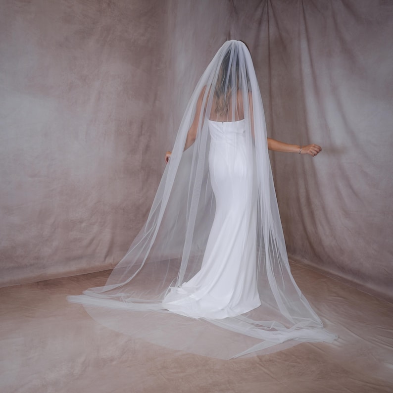May include: A long, sheer white bridal veil made of fine tulle fabric. The veil is draped over the head and extends down the back, flowing onto the floor in a wide, translucent train. The material is lightweight, semi-transparent, and smooth.
