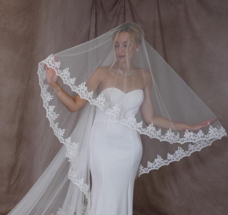 K&ouml;nnte beinhalten: Ein wei&szlig;er Brautschleier mit zarter Spitzenborte. Der transparente Schleier wird mit einem tr&auml;gerlosen wei&szlig;en Brautkleid kombiniert. Die Spitzenborte verleiht dem Brautensemble Eleganz.