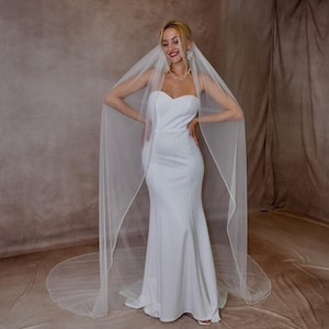 May include: A floor-length white wedding dress with a sweetheart neckline and a fitted bodice that flares at the skirt. A long, sheer white veil with a delicate finished edge drapes over the shoulders and extends onto the floor behind the dress.