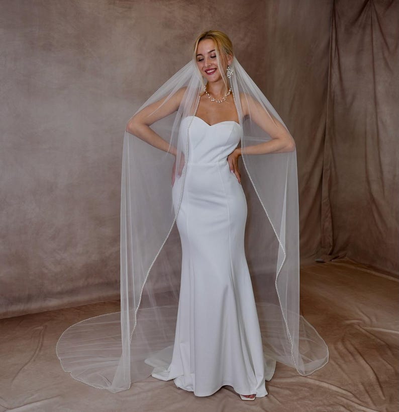 May include: A floor-length white wedding dress with a sweetheart neckline and a fitted bodice that flares at the skirt. A long, sheer white veil with a delicate finished edge drapes over the shoulders and extends onto the floor behind the dress.