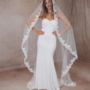 May include: A white bridal gown with a sweetheart neckline and a mermaid style. The bride is wearing a long, sheer veil with a delicate lace trim. The veil flows down the back, creating an elegant and romantic appearance.