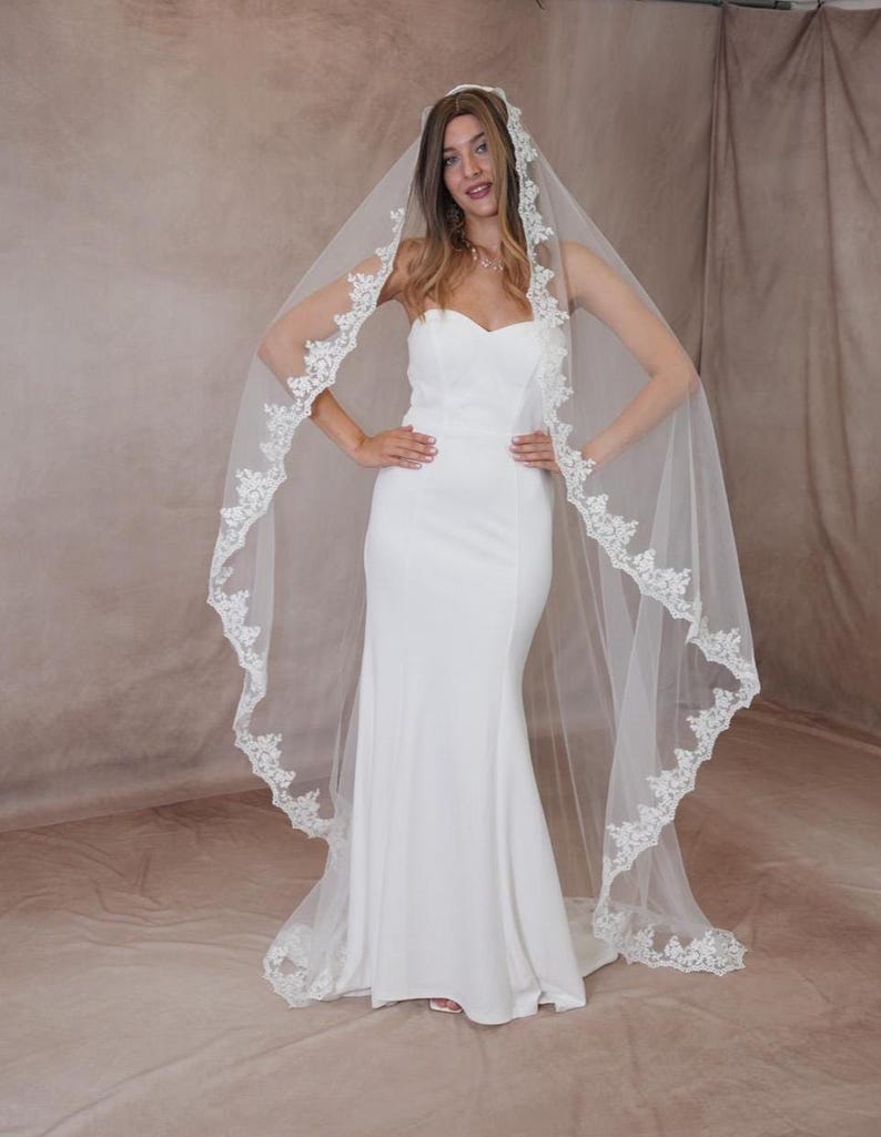 May include: A white bridal gown with a sweetheart neckline and a mermaid style. The bride is wearing a long, sheer veil with a delicate lace trim. The veil flows down the back, creating an elegant and romantic appearance.
