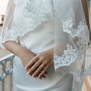 K&ouml;nnte beinhalten: Wei&szlig;es Brautkleid mit passendem Schleier. Der Schleier hat einen zarten Spitzensaum. Die H&auml;nde der Braut sind gefaltet, und sie tr&auml;gt einen Ring. Das Kleid ist ein schlichtes, elegantes Design.