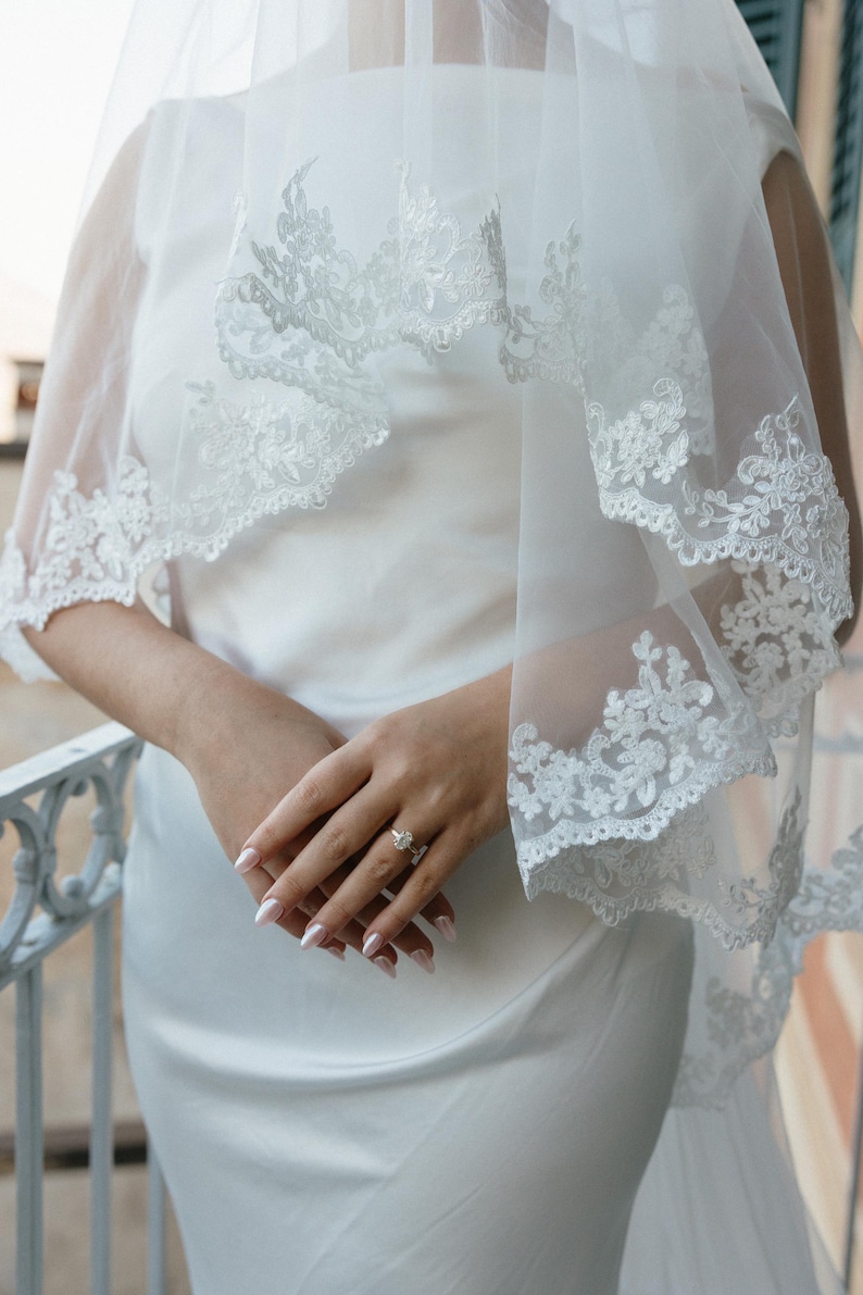 K&ouml;nnte beinhalten: Wei&szlig;es Brautkleid mit passendem Schleier. Der Schleier hat einen zarten Spitzensaum. Die H&auml;nde der Braut sind gefaltet, und sie tr&auml;gt einen Ring. Das Kleid ist ein schlichtes, elegantes Design.