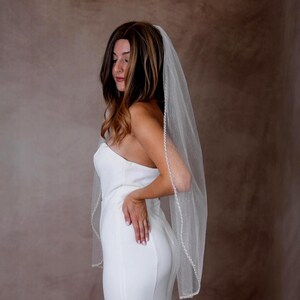 May include: A sheer white bridal veil featuring a delicate beaded trim along the edges and a subtle shimmering glitter finish throughout the tulle fabric. The veil is worn over a strapless white gown, cascading down the back in a soft, flowing drape.