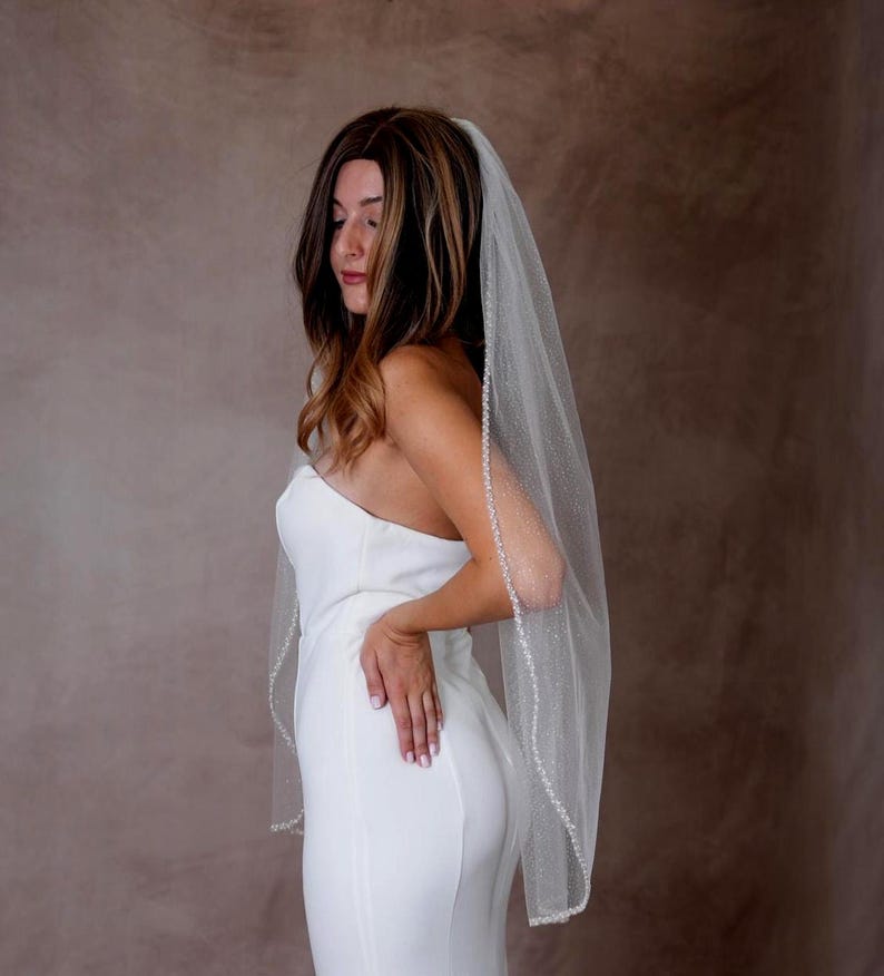 May include: A sheer white bridal veil featuring a delicate beaded trim along the edges and a subtle shimmering glitter finish throughout the tulle fabric. The veil is worn over a strapless white gown, cascading down the back in a soft, flowing drape.