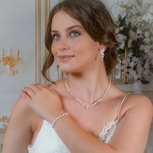 May include: A bridal jewelry set with a delicate necklace, bracelet, and earrings. The set features a sparkling crystal design with a floral motif. The jewelry is silver-toned and has a classic, elegant style.