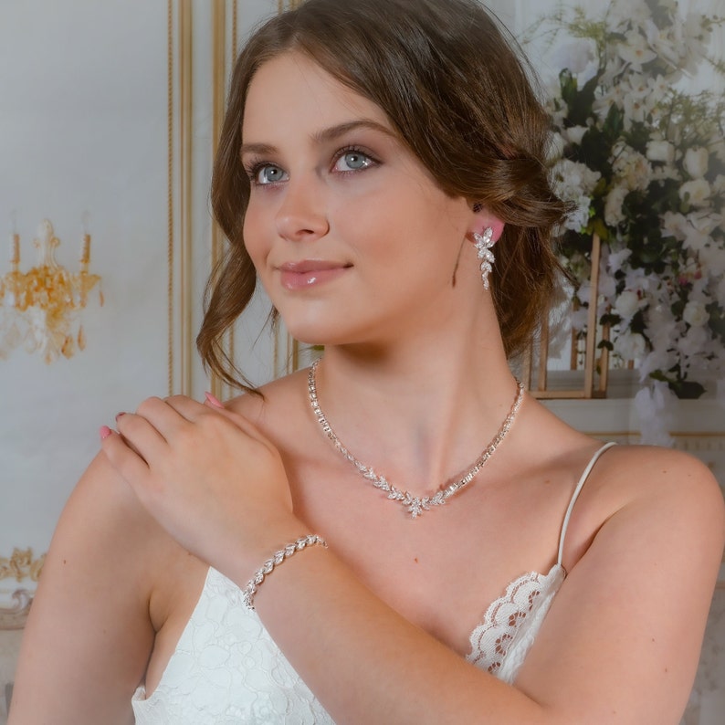 May include: A bridal jewelry set with a delicate necklace, bracelet, and earrings. The set features a sparkling crystal design with a floral motif. The jewelry is silver-toned and has a classic, elegant style.