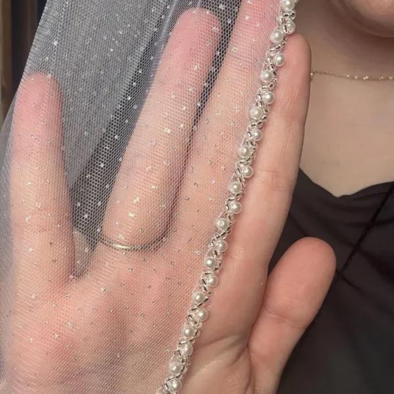 May include: A sheer white tulle veil featuring a fine glitter finish and a decorative border edge. The border is embellished with a continuous row of small, round, white pearls woven into a silver-toned metallic thread structure.