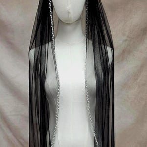Puede incluir: Un velo negro transparente drapeado sobre un maniqu&iacute;, con un borde delicado de peque&ntilde;as perlas blancas a lo largo de todo el contorno. La tela de tul ligera cuelga verticalmente, creando un efecto de capas con las perlas al frente.