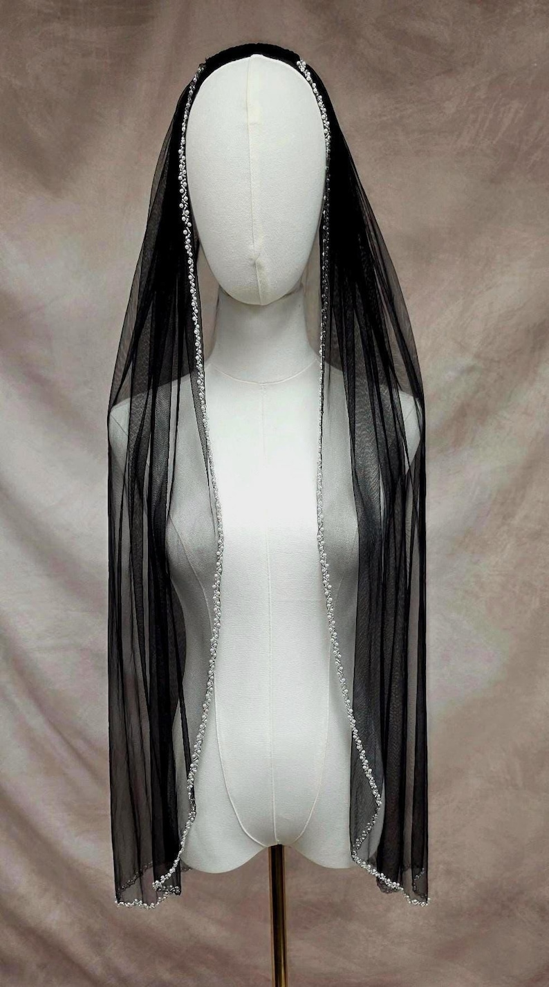 Puede incluir: Un velo negro transparente drapeado sobre un maniqu&iacute;, con un borde delicado de peque&ntilde;as perlas blancas a lo largo de todo el contorno. La tela de tul ligera cuelga verticalmente, creando un efecto de capas con las perlas al frente.