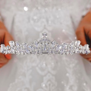 May include: A silver-toned tiara featuring a symmetrical arrangement of clear, faceted marquise and round-cut crystals. The headpiece displays a central elevated peak with smaller floral-inspired clusters extending outward along the band.