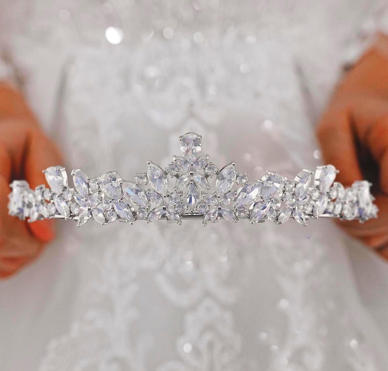 May include: A silver-toned tiara featuring a symmetrical arrangement of clear, faceted marquise and round-cut crystals. The headpiece displays a central elevated peak with smaller floral-inspired clusters extending outward along the band.