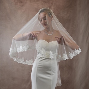K&ouml;nnte beinhalten: Eine Braut in einem wei&szlig;en, tr&auml;gerlosen Brautkleid und einem transparenten Schleier mit Spitzenbesatz. Die Braut tr&auml;gt eine Halskette und ein Armband. Der Hintergrund ist eine neutrale Farbe. Der Schleier verleiht dem Brautlook Eleganz.