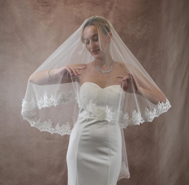 K&ouml;nnte beinhalten: Eine Braut in einem wei&szlig;en, tr&auml;gerlosen Brautkleid und einem transparenten Schleier mit Spitzenbesatz. Die Braut tr&auml;gt eine Halskette und ein Armband. Der Hintergrund ist eine neutrale Farbe. Der Schleier verleiht dem Brautlook Eleganz.