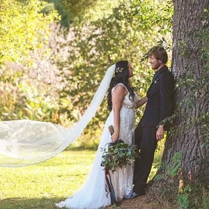Puede incluir: Una novia y un novio se encuentran cerca uno del otro en un campo de hierba, el velo de la novia ondeando en el viento. El novio se apoya en un &aacute;rbol, y la novia sostiene un ramo de vegetaci&oacute;n.