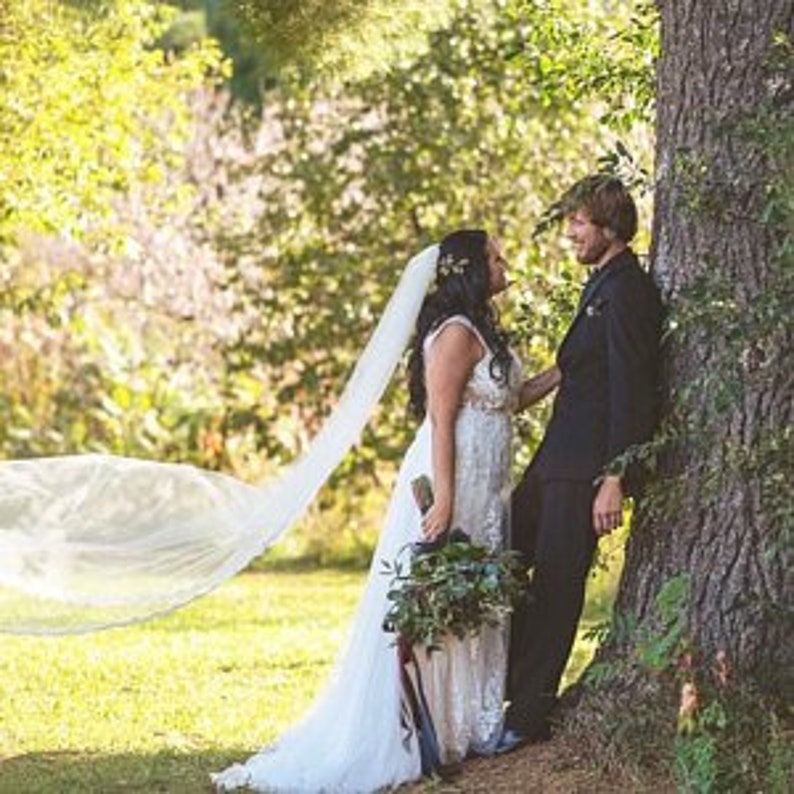 Puede incluir: Una novia y un novio se encuentran cerca uno del otro en un campo de hierba, el velo de la novia ondeando en el viento. El novio se apoya en un &aacute;rbol, y la novia sostiene un ramo de vegetaci&oacute;n.