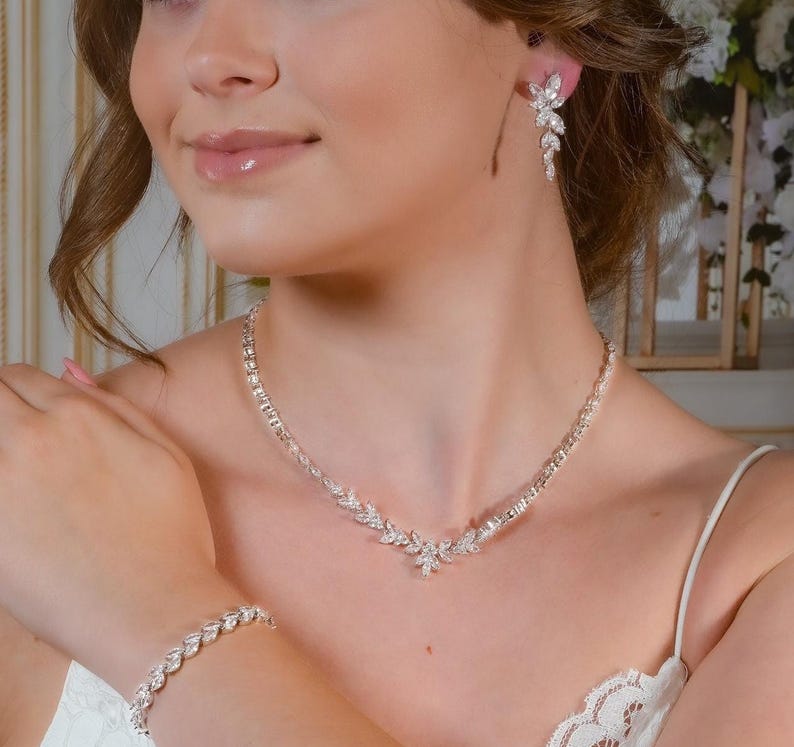 May include: A sparkling jewelry set featuring a necklace, bracelet, and earrings. The necklace and bracelet have a leaf-like design, and the earrings dangle. The jewelry is silver-toned and appears to be made of crystals or rhinestones.