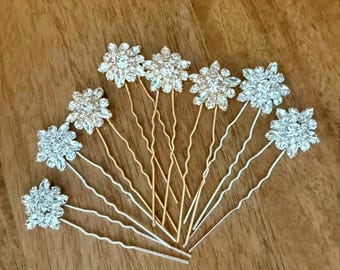 May include: A set of eight hair pins featuring floral-shaped embellishments covered in clear, sparkling crystals. The pins are arranged in a fan shape, with some pins featuring gold-toned metal stems and others featuring silver-toned metal stems.