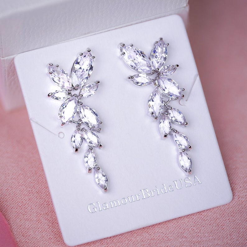 May include: A pair of silver dangle earrings with clear crystal stones in a floral design. The earrings are displayed on a white card with the text "GlamourBrideUSA".