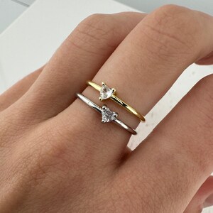 May include: Two dainty rings with heart-shaped clear stones. The ring on top is gold, and the ring on the bottom is silver.