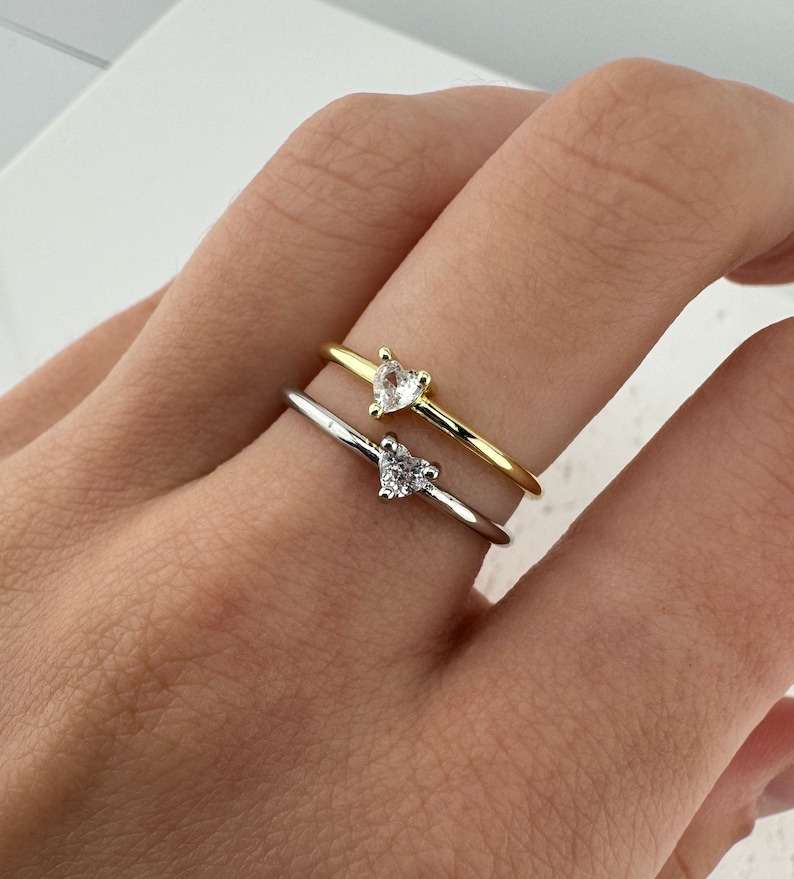 May include: Two dainty rings with heart-shaped clear stones. The ring on top is gold, and the ring on the bottom is silver.