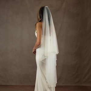 May include: A two-tier bridal veil made of sheer white tulle, featuring a delicate beaded edge along the hem. The veil is attached to a hair comb and drapes down the back, layered over a simple, floor-length white sleeveless gown.