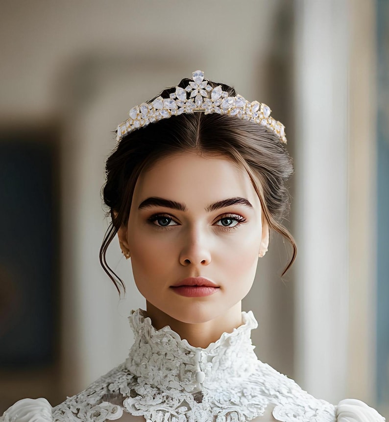 May include: A gold-toned tiara featuring a symmetrical arrangement of clear, pear-shaped and marquise-cut crystals. The headpiece displays a central floral motif with radiating crystal petals, set against a metallic frame that rests on the head.