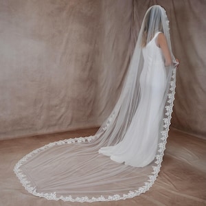 K&ouml;nnte beinhalten: Ein langer, wei&szlig;er Brautschleier mit zarter Spitzenborte. Der Schleier ist &uuml;ber einem wei&szlig;en Kleid drapiert und erzeugt einen flie&szlig;enden, eleganten Look. Die Spitzenborte verleiht dem Brautaccessoire einen Hauch von Raffinesse.